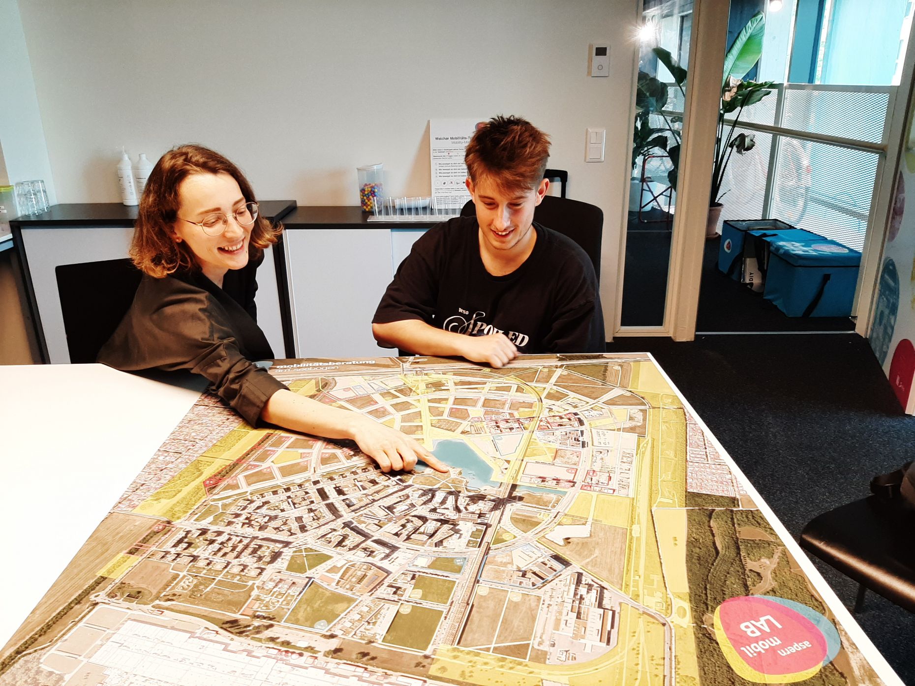 Magdalena Bürbaumer und Marco Farinelli sitzen mit einer Karte der Seestadt an einem Tisch im Büro des aspern.mobil Labs. Magdalena deutet mit dem Zeigefinger auf ein Gebiet in der Seestadt.